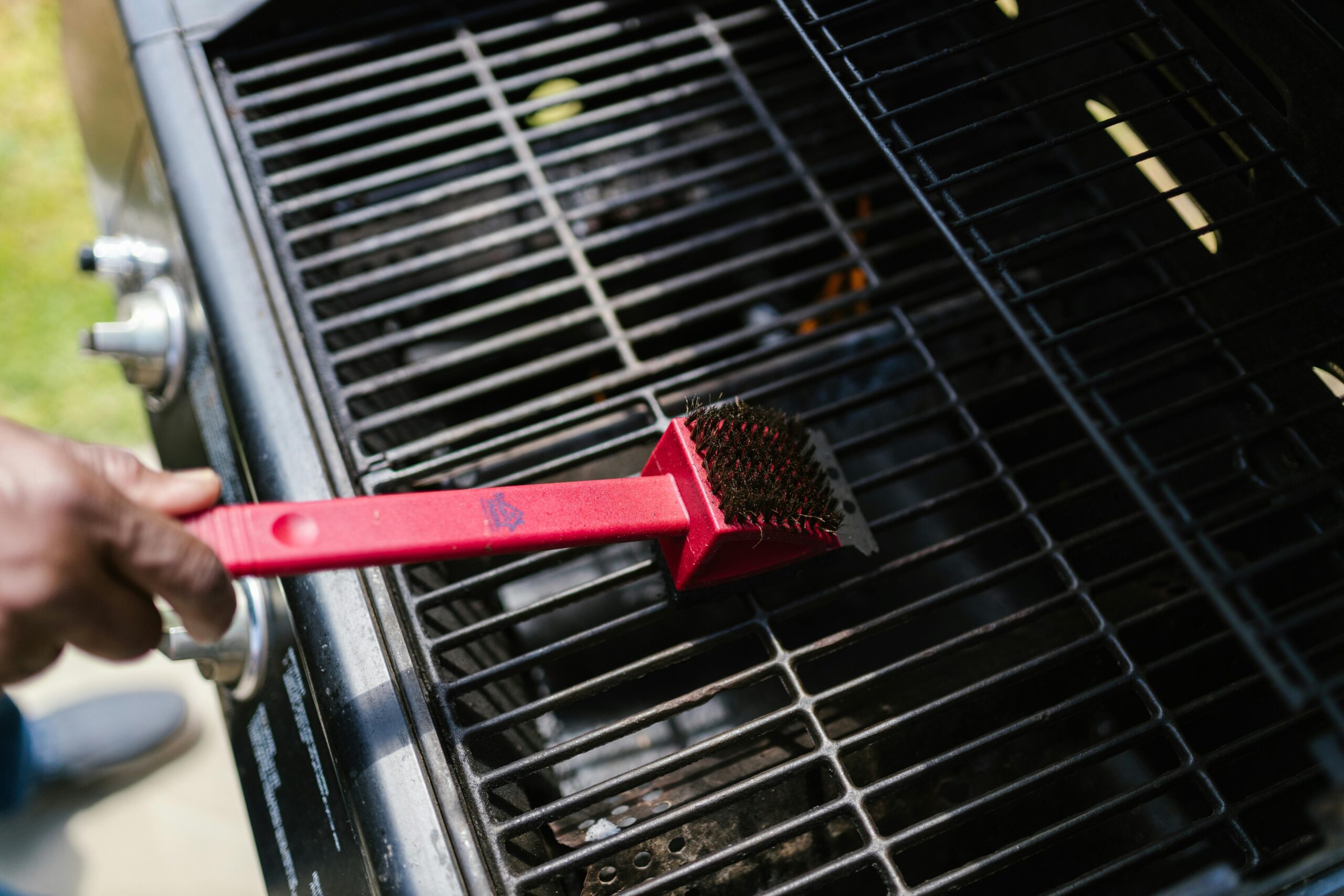 A person cleaning a barbecue grill with a red brush in daylight. Ideal for BBQ enthusiasts.